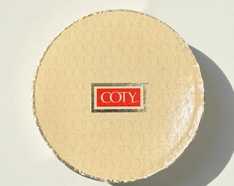 May include: A round, beige Coty compact with gold trim. The center features a red rectangle with the word "COTY" in white lettering. The background is patterned with the word "COTY".