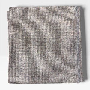Puede incluir: Un cuadrado doblado de tela tejida gris y blanca. La tela tiene un patrón pequeño y repetitivo, creando una apariencia texturizada. Los bordes de la tela están cuidadosamente doblados, lo que sugiere un producto terminado, posiblemente un artículo textil.