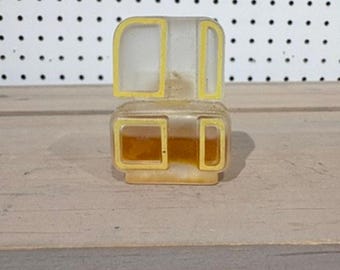 Op de afbeelding: Een vintage, doorzichtige plastic container met gele rand. De container heeft een rechthoekige basis en een afgerond bovendeel. De binnenkant lijkt een vloeistof te bevatten. De container staat op een houten oppervlak.