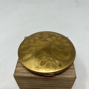 Op de afbeelding: Een gouden compact met een bloemenmotief. De compact staat op een houten blok.