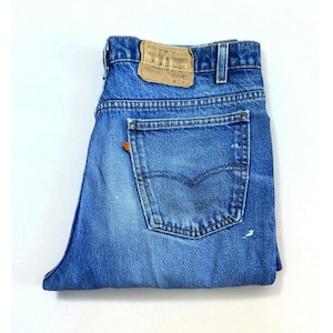 May include: Blue denim jeans featuring a tan leather patch on the back waistband with Levi Strauss & Co. text. The jeans include a single back pocket with an orange tab on the side and a faded wash with light distressing on the fabric surface.