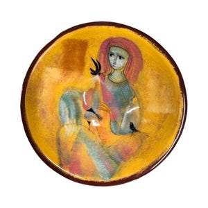 Pode incluir: Uma tigela de cerâmica redonda com um fundo amarelo vibrante e uma ilustração pintada de uma figura com cabelo avermelhado segurando um pequeno pássaro preto. A tigela tem uma borda castanho-escura e um acabamento texturizado e pictórico.