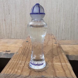 Pode incluir: Um frasco de perfume de vidro transparente com uma tampa azul e branca. O frasco tem uma forma esguia de ampulheta e é preenchido com um líquido transparente. A base do frasco tem o texto "more muss mimmina".