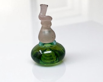May include: A small, antique glass perfume bottle. The bottle has a round, green base with a clear, spiraled top. The base is a deep green color, and the top is a frosted, pale pink. The bottle is sitting on a white surface.