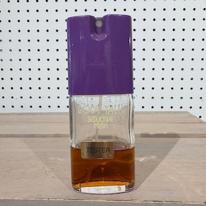 May include: A clear glass perfume bottle with a purple cap. The bottle contains a brown liquid and has the words "BODY TALK Seductive Musk" printed on it. A gold "TESTER" label is affixed to the front.
