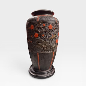 May include: A dark brown vase with a detailed relief design. The vase features red accents and a carved pattern of flowers, branches, and a bird. The vase sits on a round base.