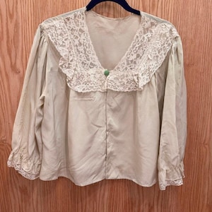 May include: A beige long-sleeve blouse featuring a large, wide lace collar with floral patterns, a single small green button at the neckline, and matching lace trim at the cuffs. The garment has a loose, boxy fit and a V-neckline.