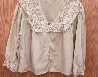 Op de afbeelding: Een beige blouse met lange mouwen, voorzien van een grote, brede kanten kraag met bloemmotieven, een enkele kleine groene knoop bij de halslijn en bijpassende kanten randen aan de manchetten. Het kledingstuk heeft een losse pasvorm en een V-hals.