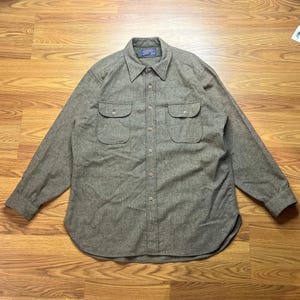 Puede incluir: Una camisa de manga larga con botones en un color marrón topo jaspeado. La camisa presenta un cuello en punta, dos bolsillos en el pecho con solapas abotonadas y un cierre frontal de botones completo. La tela tiene un tejido texturizado visible.