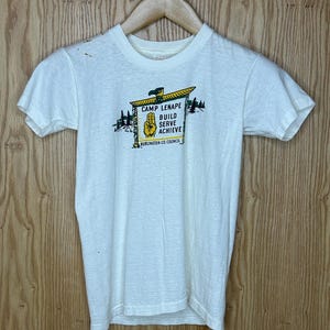 May include: Off-white t-shirt with a graphic featuring the text "CAMP LENAPE BUILD SERVE ACHIEVE" and a hand symbol. The graphic is in yellow, green, and black. The shirt is hanging on a wooden hanger.