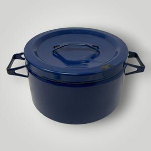 May include: A deep, round, dark blue enamelled metal cooking pot with a matching lid. The pot features two angular side handles and a central handle on the lid. There are small areas of chipped enamel visible on the lid and the pot rim.