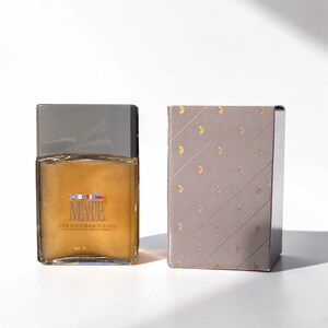 Peut inclure: Une bouteille de verre rectangulaire de baume après-rasage avec un bouchon argenté et une boîte assortie. La bouteille porte une étiquette avec le mot "MATE" et un motif de drapeau. La boîte est grise avec un motif doré.