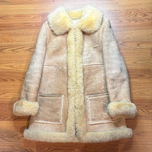 May include: A beige shearling coat featuring a thick, cream-colored fur collar, cuffs, and hem. The coat has a smooth, tan suede-like exterior with visible paneling, two large front patch pockets, and a long, central fur-lined front opening.