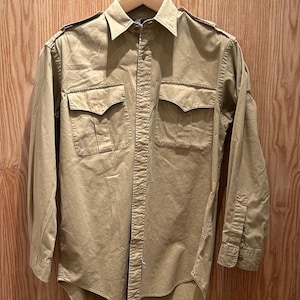 Puede incluir: Una camisa abotonada de color beige con dos bolsillos en el pecho y charreteras en los hombros. La camisa de manga larga está colgada en una percha de madera sobre un fondo de madera.
