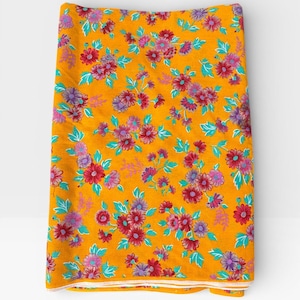 Op de afbeelding: Gevouwen stof met een levendig bloemenpatroon. Het ontwerp toont clusters van rode, paarse en roze bloemen met turquoise bladeren op een goudgele achtergrond. De randen zijn afgewerkt met een witte rand.