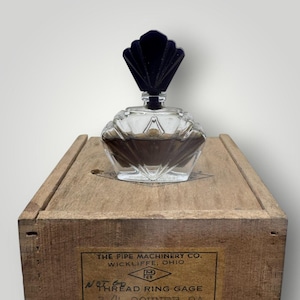 May include: A vintage perfume bottle with a clear, faceted glass body and a dark blue, fan-shaped stopper. The bottle sits on a wooden box with a label from The Pipe Machinery Co. in Wickliffe, Ohio.