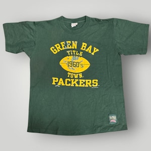 Puede incluir: Camiseta verde oscuro con texto amarillo "GREEN BAY TITLE 1960'S TOWN PACKERS". La camiseta tiene cuello redondo y mangas cortas. Una pequeña etiqueta es visible en la parte inferior. Esta camiseta vintage es un objeto de colección deportiva.