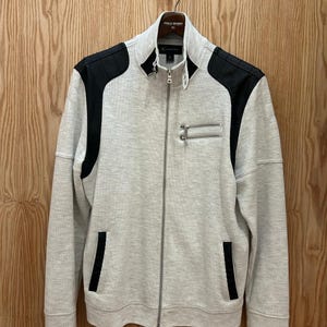 May include: A light gray zip-up jacket with black shoulder patches and pocket accents. The jacket has a high collar and two zippered chest pockets. The jacket is hanging on a wooden background.