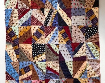 Op de afbeelding: Een patchwork quilt met een geometrisch ontwerp. De quilt heeft een verscheidenheid aan stoffen in tinten blauw, bruin, rood en crème. De quilt is gemaakt van vierkanten en driehoeken, waardoor een uniek en kleurrijk patroon ontstaat.