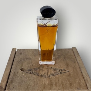Puede incluir: Un frasco de perfume vintage con una tapa negra y un cuerpo de vidrio transparente y rectangular lleno de líquido ámbar. El frasco se encuentra sobre una superficie de madera con un logotipo en forma de diamante que dice "THE PIPE MACHINERY CO. CLEVELAND OHIO U.S.A."
