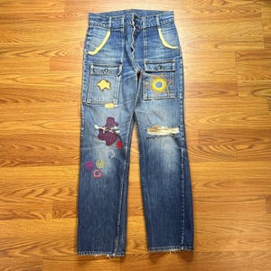 May include: Blue denim jeans featuring a high-waisted fit, two large front pockets with yellow trim, and a button fly. The legs are decorated with various colorful embroidered shapes, including a sun, circles, and a purple patch, plus a distressed rip.