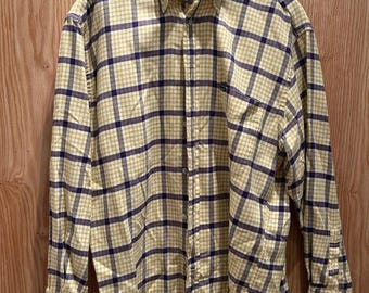 Op de afbeelding: Een lange mouwen overhemd met een geel en marineblauw ruitpatroon. Het overhemd hangt aan een houten hanger tegen een houten achtergrond. Het overhemd heeft een button-down kraag en een borstzak.