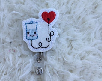 May include: A white glitter badge reel with a blue and white cartoon IV bag holding a red heart balloon. The IV bag has a smiling face.