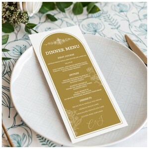May include: A gold-colored dinner menu card with floral accents and the text "DINNER MENU." The menu lists courses such as salad, entree, and desserts. The card is placed on a white plate with a knife.