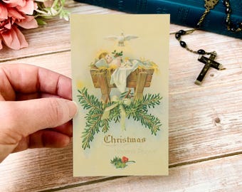 Op de afbeelding: Een vintage kerstkaart met een kerststal met een baby in een kribbe, een duif en de tekst "Christmas Novena Prayer". De kaart is versierd met groen en een strik.
