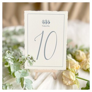 May include: A cream-colored table number card with a light blue border and the number 10 in a matching script font. The card is displayed on a small wooden easel, with floral accents.