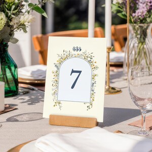 May include: A cream-colored table number card with the number 7 in navy blue, framed by a floral arch design. The card is held in place by a small wooden stand, with flowers and candles in the background, suggesting a wedding or event setting.