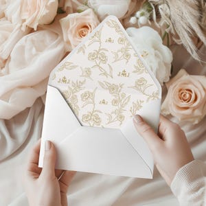 Puede incluir: Un sobre blanco se abre, revelando un interior con estampado floral. El diseño interior presenta rosas y follaje dorados sobre un fondo crema. El sobre está rodeado de flores desenfocadas, lo que sugiere una boda o una ocasión especial.