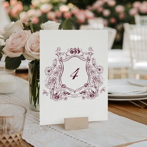 May include: A cream-colored table number card with a burgundy floral crest design and the number 4. The crest includes the names "Elizabeth" and "Colten" and the word "Durrer". The card is held by a small wooden stand, with a bouquet of pink roses in the background.