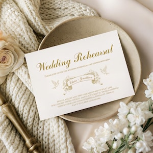 May include: A wedding rehearsal invitation card with gold lettering on a cream background. The card reads "Wedding Rehearsal" and includes the names "Claire + Jonathan". The card is on a beige plate, with white flowers and a gold-colored decorative element.
