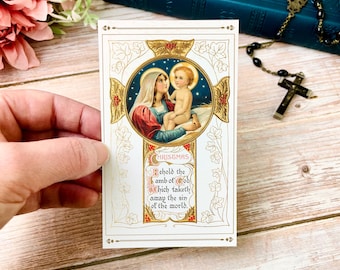 Op de afbeelding: Een vintage kerstkaart met een gouden omlijsting van een afbeelding van Maria die baby Jezus vasthoudt. De kaart heeft een witte achtergrond met gouden en rode accenten. De tekst op de kaart luidt "Kerstmis" en "Zie, het Lam Gods, dat de zonde der wereld wegneemt."