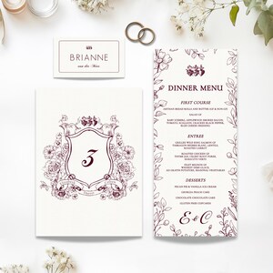 May include: Elegant wedding stationery set featuring a table number card, dinner menu, and place card. The design includes floral accents and burgundy text on a cream background. Gold wedding rings are also visible.