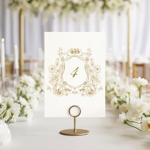 May include: A gold-colored table number card with the number 4, displayed in a gold metal holder. The card features a floral crest design with the names "Claire" and "Jonathan" on banners. The background includes white flowers and candles.