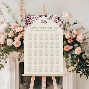 May include: A cream-colored wedding seating chart on a wooden easel. The chart features a floral border and the text "PLEASE TAKE YOUR SEAT". Names are listed in columns, with the names of the couple and the wedding date at the top. The floral design is a deep burgundy color.
