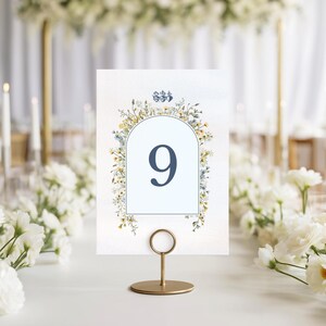 May include: A table number card with the number 9 in a navy blue font, set in a gold metal holder. The card is decorated with a floral design in yellow, blue, and green, perfect for weddings or special events.