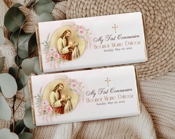 May include: Two white chocolate bar wrappers with a religious theme. Each wrapper features a picture of Jesus with a young girl, surrounded by pink flowers and a gold cross. The text on the wrapper reads "My First Communion Eleanor Marie Duleau Sunday, May 26, 2025".