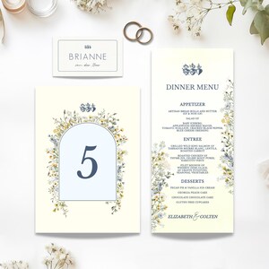 May include: Wedding stationery set featuring a table number, place card, and dinner menu. The designs have a cream background with floral accents in yellow, blue, and white. Gold wedding rings are also visible.