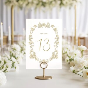 May include: A gold table number card with the number 13, displayed in a gold holder. The card features a floral wreath design and the words "TABLE NO." in gold. The background includes white flowers and candles.