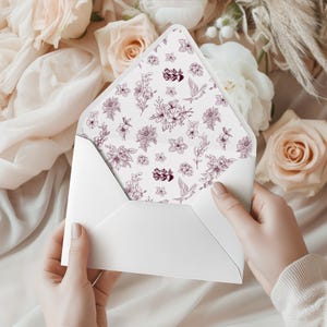 May include: A white envelope is held open, displaying a floral patterned interior. The design features a delicate arrangement of flowers and birds in shades of purple and burgundy against a white background. The envelope is surrounded by soft, light-coloured flowers.