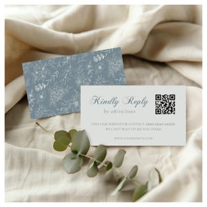 May include: Two light blue reply cards with floral designs. One card features a floral pattern, while the other has the text "Kindly Reply" and a QR code. The cards also include contact information and a website address.