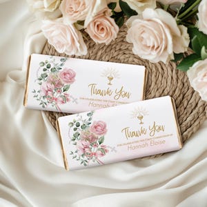 May include: Two chocolate bar wrappers with floral designs and the text "Thank You" in gold. The wrappers feature pink roses, greenery, and the name "Hannah Eloise". The bars are displayed on a woven mat with roses.