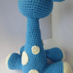 May include: A blue crocheted giraffe toy with white spots. The giraffe is sitting on a white surface.