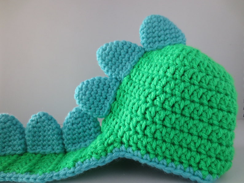 May include: Green and blue crocheted dinosaur hat with a long tail. The hat has a pointed top and a row of blue spikes along the back.