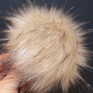 May include: A fluffy, light brown faux fur pom pom with dark brown accents. The pom pom is made of soft, synthetic fibers and is perfect for adding a touch of whimsy to hats, scarves, or other accessories.
