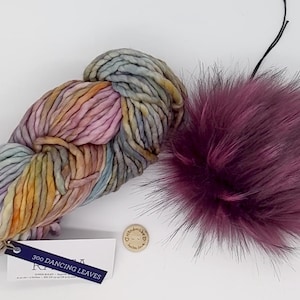 Puede incluir: Un ovillo de hilo multicolor con un esquema de color rosa, morado y azul. El hilo está etiquetado como "300 Dancing Leaves". Se incluye un pequeño pompón redondo marrón. El hilo es para tejer o hacer ganchillo.