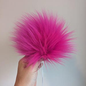 Könnte beinhalten: Ein leuchtend pinkfarbener Kunstfell-Bommel mit langen, stacheligen Fasern. Der Bommel wird von einer Hand gehalten, an der ein dünner weißer Faden befestigt ist. Der Hintergrund ist ein schlichtes, hellgraues.